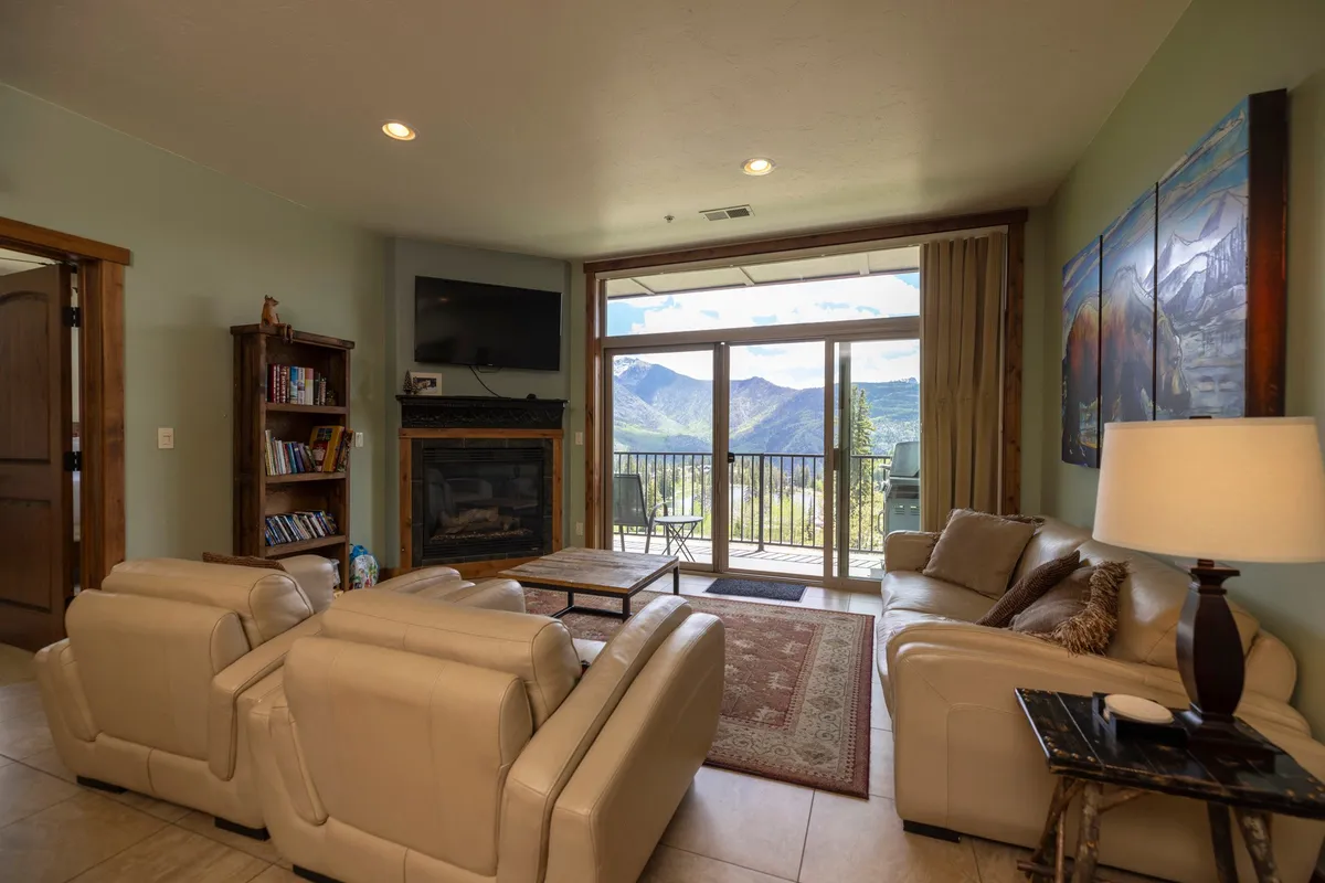 A plush living room with luxurious leather sofas, a crackling fireplace, and wide windows offering sunset views of the mountain range. Artworks and decor subtly reflect the alpine setting, while the balcony is visible, suggesting direct access to fresh mountain air.