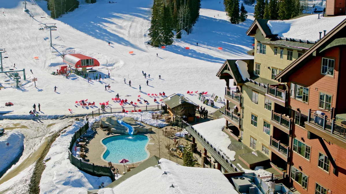 A lively scene featuring ski slopes, the pool area, a cozy lodge, and several skiers in action. The sun sets behind the mountains, bathing the area in warm light and highlighting the mountain resort's vibrant energy.