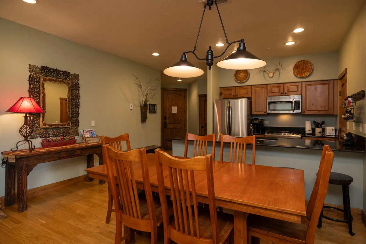 A warm, welcoming dining area featuring a spacious table, a red lamp, a decorative mirror, and an adjacent kitchen. The setting is perfect for sharing meals, entertaining guests, and creating memorable moments.