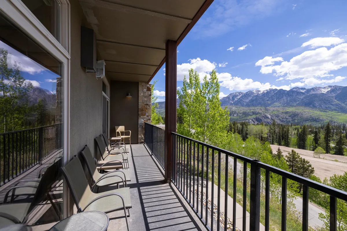 The image showcases a spacious condo balcony with comfortable seating, surrounded by lush greenery and offering unobstructed views of the majestic Needles Mountains. The scene is bathed in tranquil evening light, inviting guests to relax and enjoy nature’s beauty as the sun sets. The atmosphere is serene, with the promise of peaceful afternoons, breathtaking sunrise views, and magical stargazing at night.