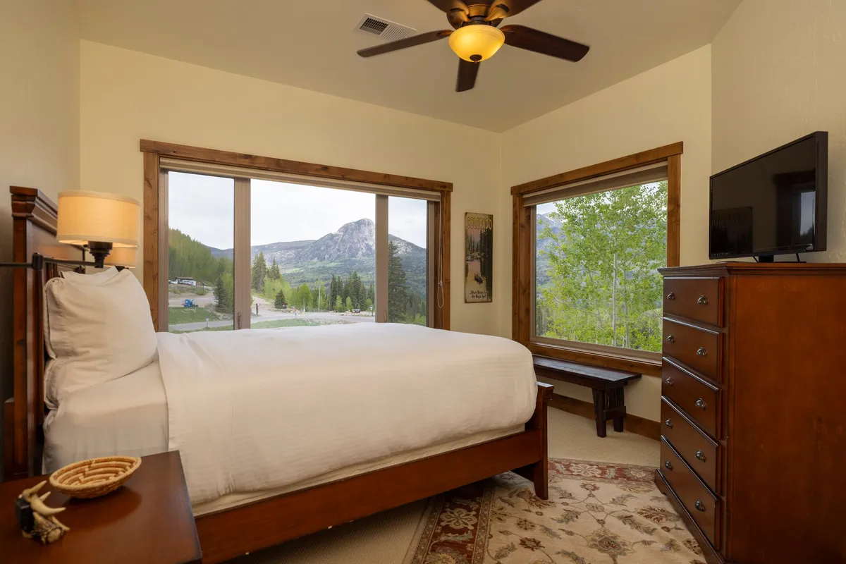 A serene bedroom scene featuring a bed with crisp white linens, large windows framing a mountain view, a wooden dresser, and a decorative lamp. The space exudes tranquility and relaxation, ideal for unwinding after a day outdoors.