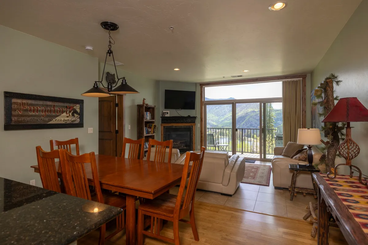 A sun-filled open living and dining space with a large sofa, elegant dining table, and expansive windows framing majestic mountain views. A stone fireplace anchors the seating area, while the setting sun casts a golden glow across the room, enhancing the warm and welcoming mood.