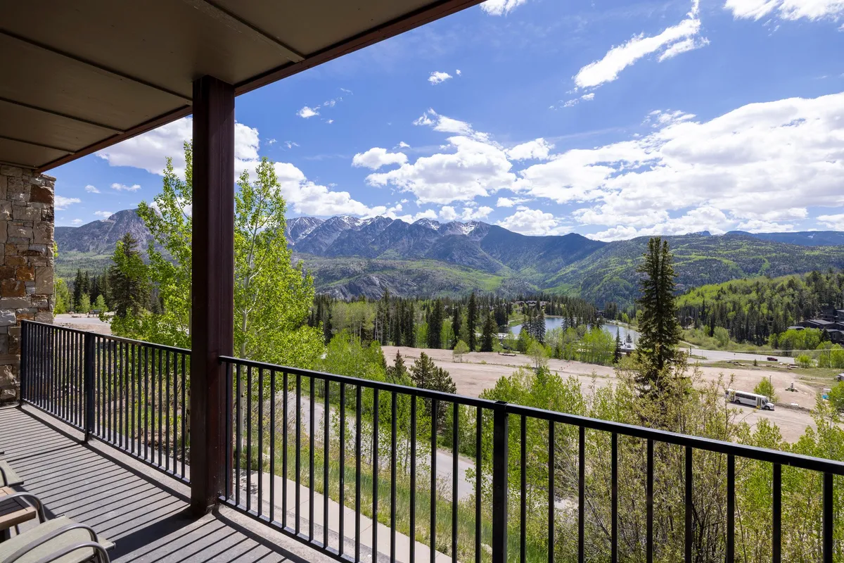 A serene view from a condo balcony looks out over a sunlit mountain range, with wild greenery below and a vivid blue sky. The setting is perfect for relaxing after a day of exploration, capturing moments of reflection or sunset-viewing with family from your private home base.