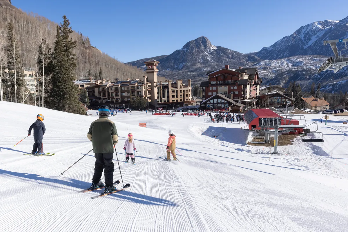 This image captures skiers on a gentle slope with a dramatic mountain backdrop and the ski resort buildings visible below. Families and groups are seen enjoying the snowy terrain together, suggesting both adventure and togetherness. The scene is bright and joyful, highlighting winter outdoor activities accessible directly from the resort.