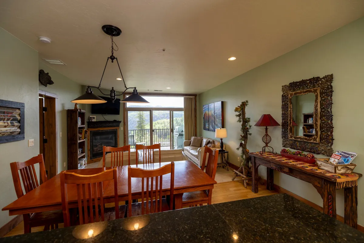 The image shows a stylish and cozy living/dining space featuring a dining table, plush sofa, and a large window flooding the room with natural light. Subtle décor touches and the window view suggest proximity to the slopes and plaza.