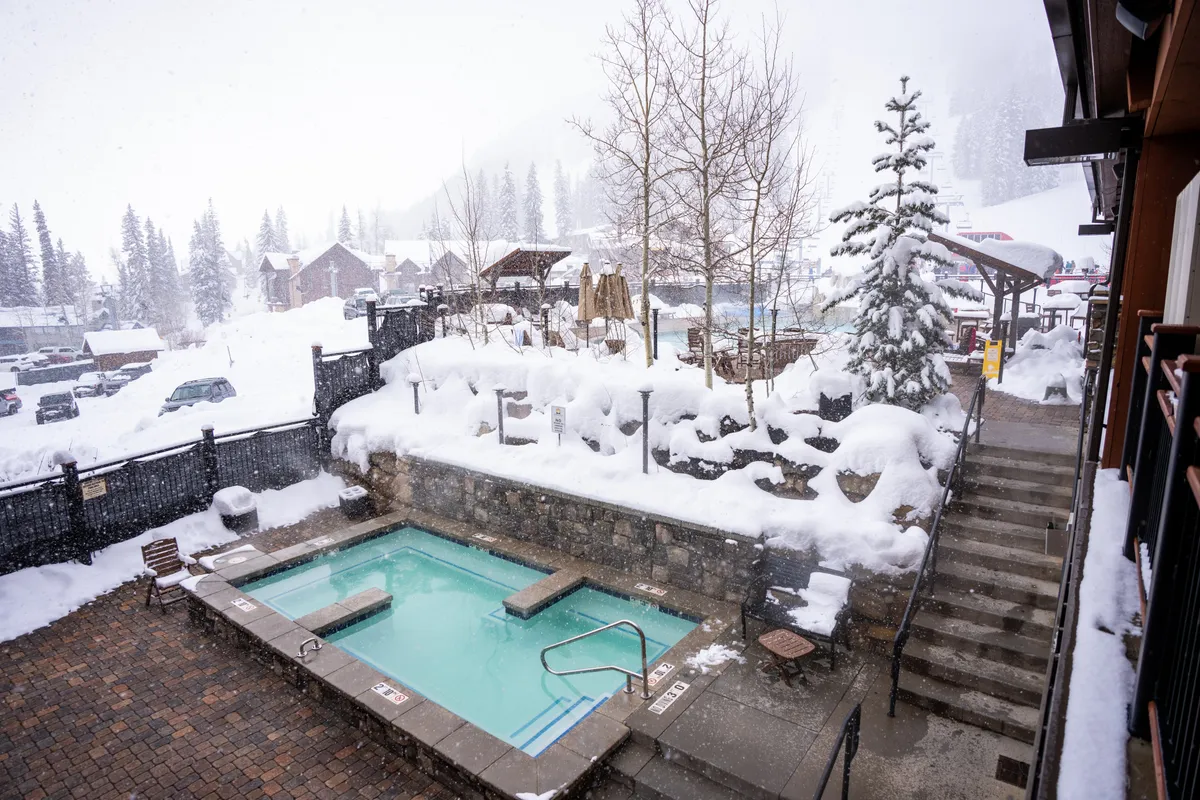 A steaming hot tub sits surrounded by snow and trees, offering a tranquil spot to relax after a day on the mountain. The winter landscape adds to the sense of exclusive, cozy retreat.