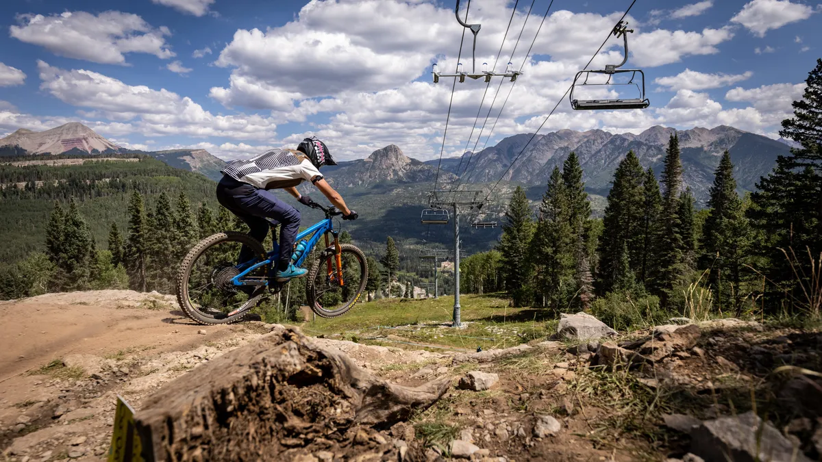 A dynamic summer scene shows a mountain biker descending a trail near a chairlift, with lush forests and towering mountains in the background. The image conveys excitement and the range of summer activities available, from biking and hiking to scenic chairlift rides, all in close proximity to the resort.